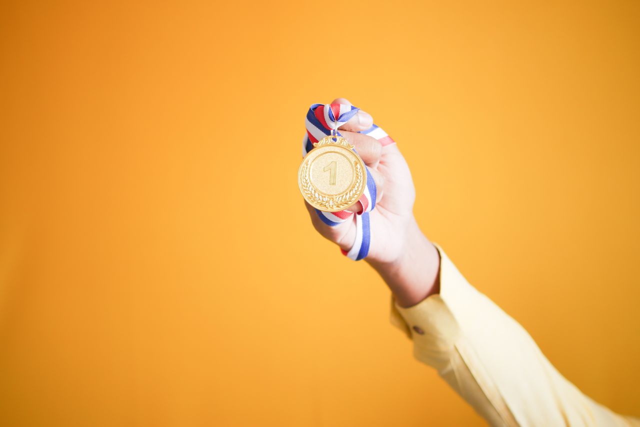 Close-up of hand holding a gold medal with a red, white, and blue ribbon against a vibrant orange background.